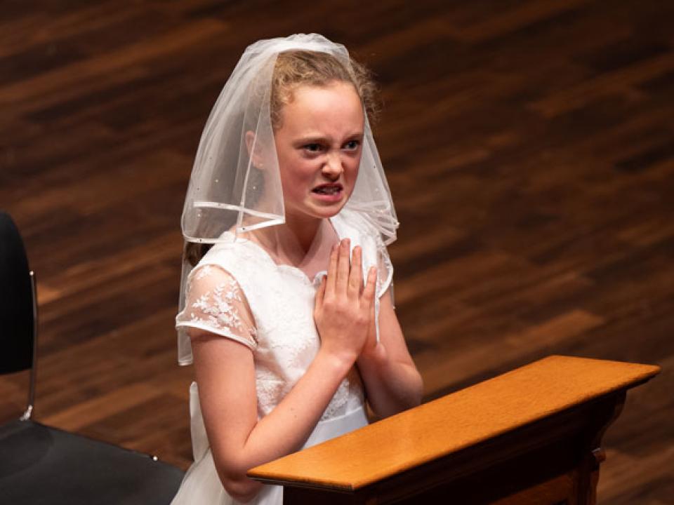 A RIAM drama student dressed in a communion dress and kneeling in prayer-like fashion performs an excerpt from Hail Mary by Eoin Colfer
