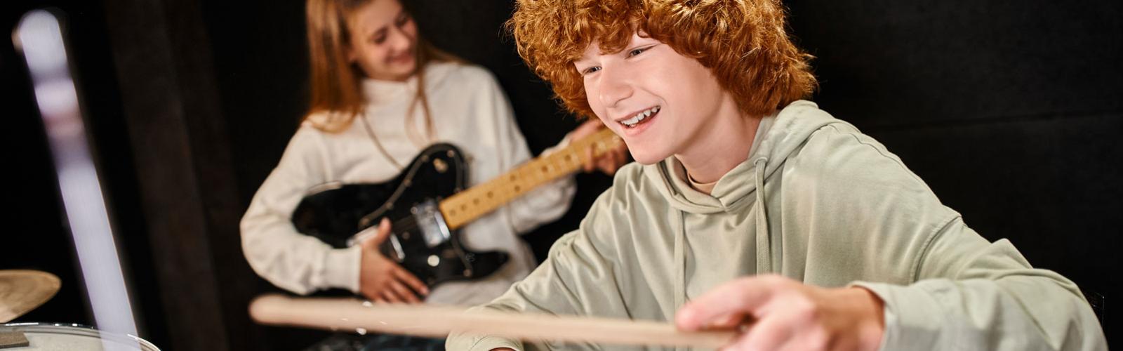 A joyful teenage boy with a mop of red hair in casual attire playing drums next to a young female guitarist with long blond hair